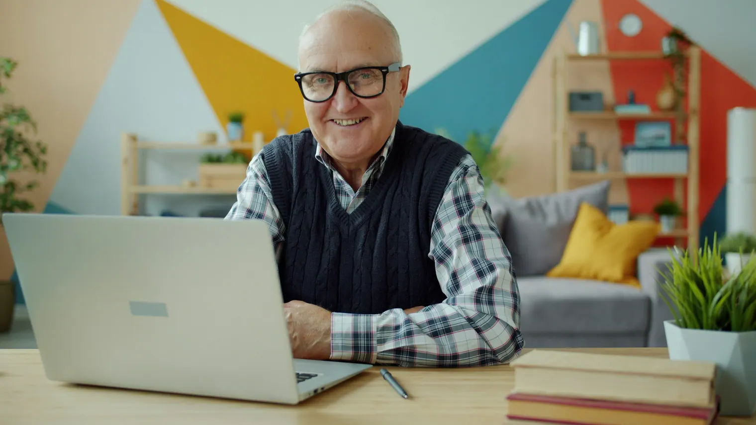 A smiling older man in glasses and a sweater vest sitting at a laptop in a brightly painted living room